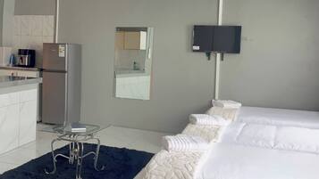 A hotel room with a kitchenette, a bed with white bedding, a small table, and a flat-screen TV mounted on the wall.