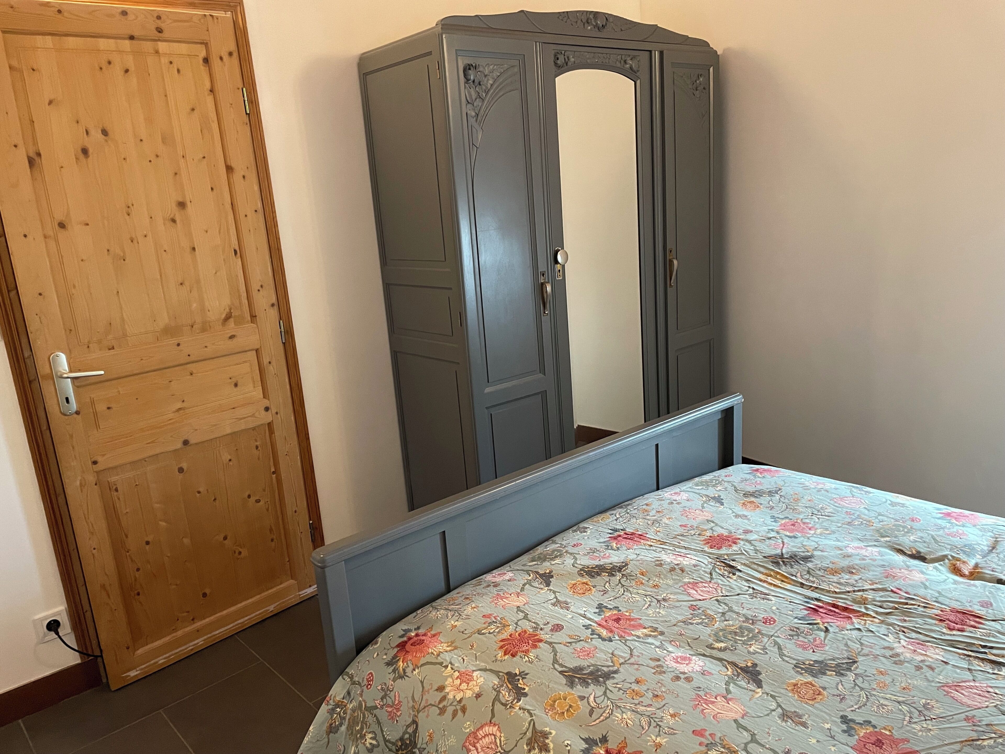 A bedroom with a wooden door, a grey wardrobe, and a bed with a floral bedspread.