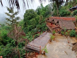 Un pont en bois traditionnel mène à un bâtiment à toit de chaume, niché au cœur d’une végétation luxuriante.