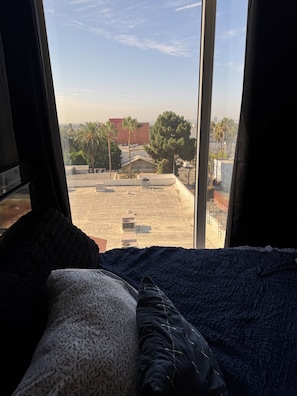 A bed with a patterned pillow and a dark blue blanket, a window with a view of a rooftop and buildings, and a clear sky. - Modern Studio with balcony in the center of Hollywood Walk of Fame area (Los Angeles)