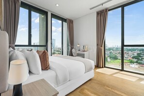 A modern bedroom with a large bed, a desk, and a view of a cityscape through large windows. - Arova by Kozystay - Cilandak (South Jakarta)