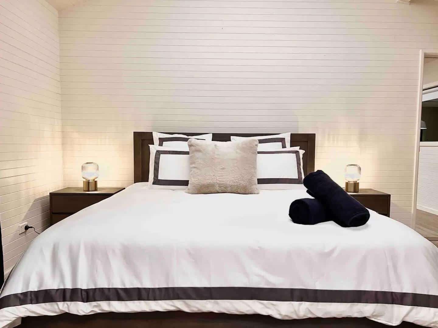 Una cama bien hecha con colcha a rayas blancas y oscuras, un almohadón mullido y dos lámparas de noche sobre mesitas de madera.
