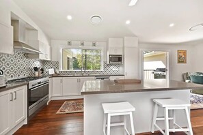 A modern kitchen with white cabinetry, a large island with bar stools, a microwave, and a window with a view of the outdoors. - Laguna Homestead - Scenic Country Escape (Cessnock City Council)