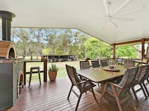 An outdoor patio with a wooden table and chairs, a ceiling fan, and a brick oven.