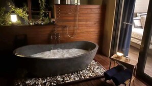 A large stone bathtub with a wooden deck, a small table with a lamp, and a window with curtains. - Laguna  Farmhouse Studio (Laguna)