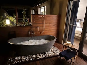 A large stone bathtub with a wooden deck, a small table with a lamp, and a window with curtains.