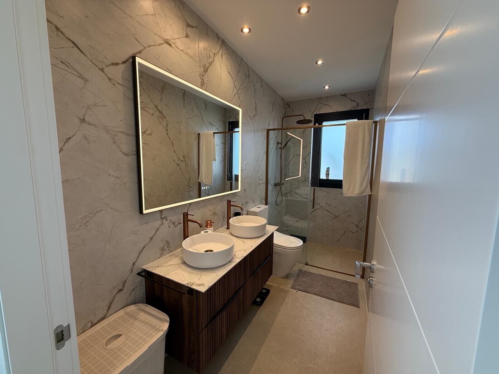 Un baño moderno con pared de mármol, un espejo grande, dos lavamanos, una ducha y un mueble de tocador de madera.
