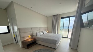 A modern bedroom with a large bed, a nightstand, a round rug, and a view of the sea through the window.