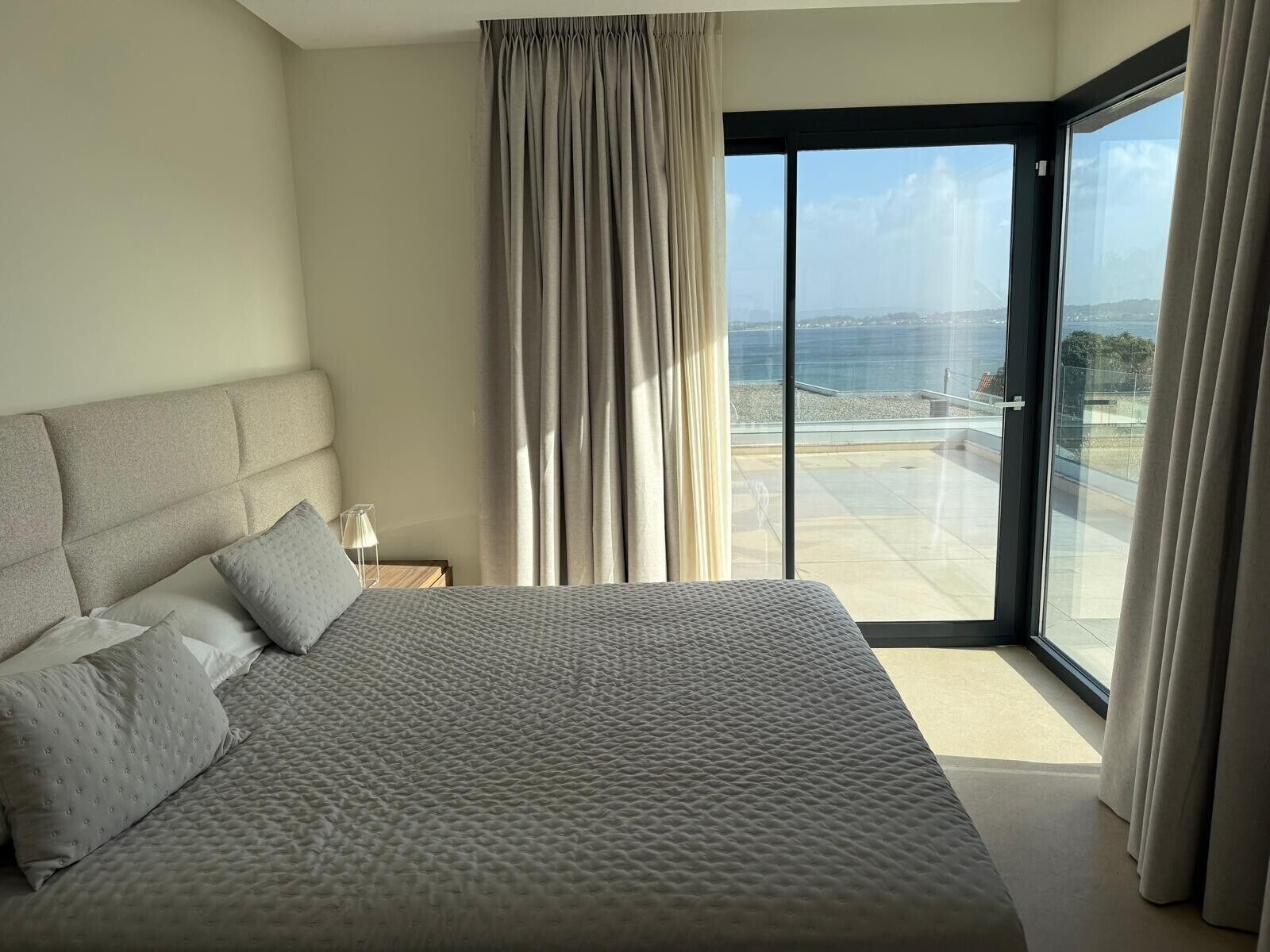 A bedroom with a large bed, a nightstand, a lamp, and a view of the sea through a sliding glass door.