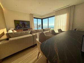 A modern hotel room with a large sofa, a dining table, and a TV mounted on the wall.