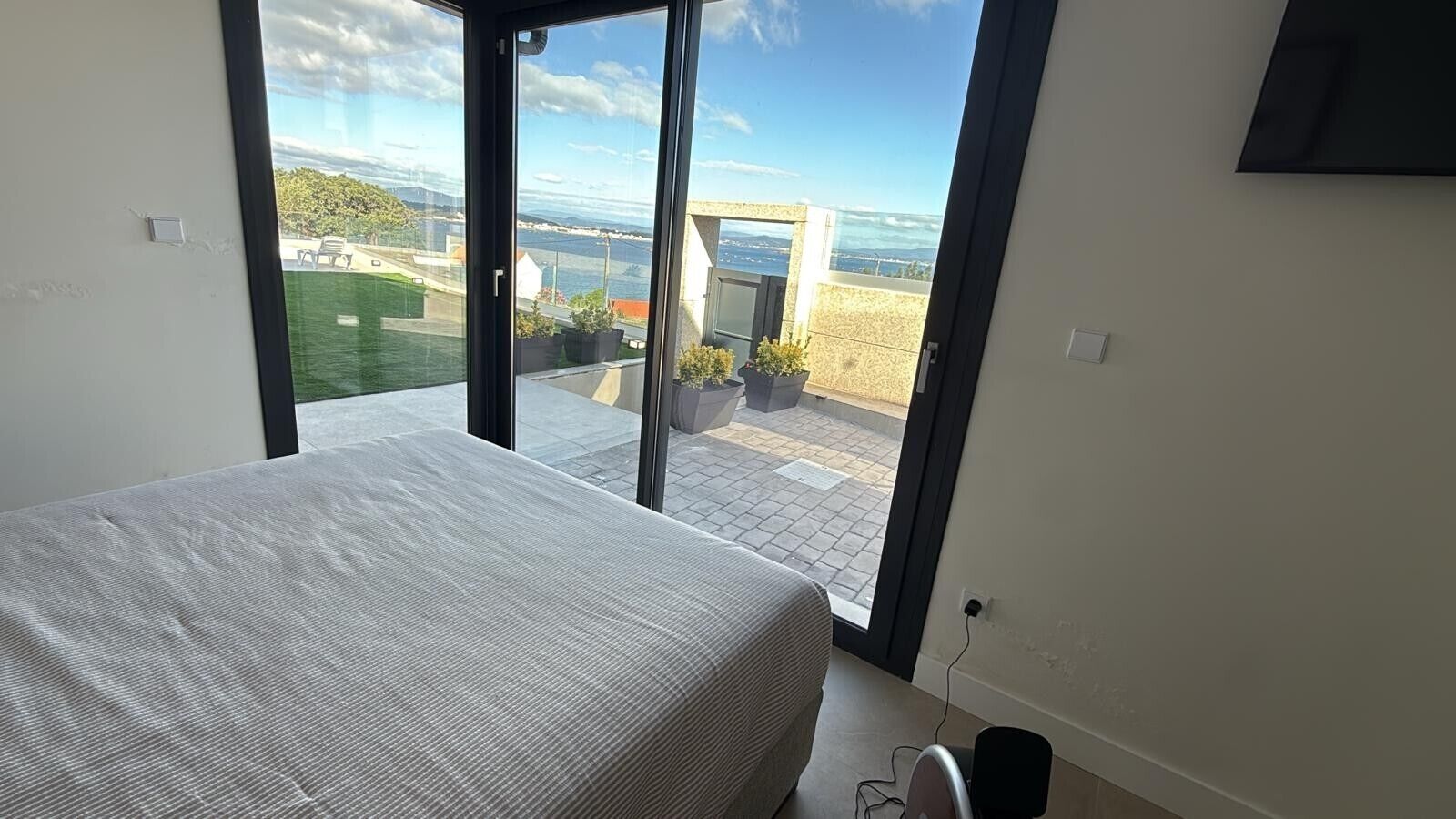 A bedroom with a bed, a sliding glass door leading to a balcony with a view of the sea and greenery.