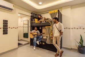 Habitación con literas, una persona tocando guitarra, otra de pie y un tercero sentado en la litera inferior.