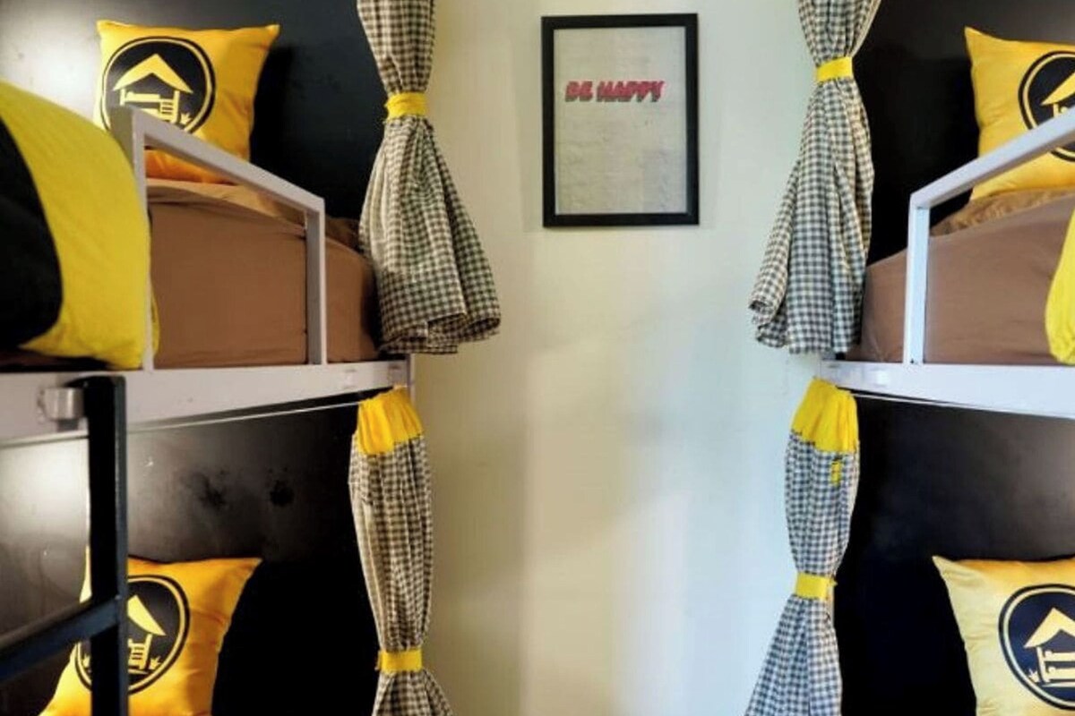Een slaapkamer met stapelbedden, geruite gordijnen en gele kussens met een logo van een huis.