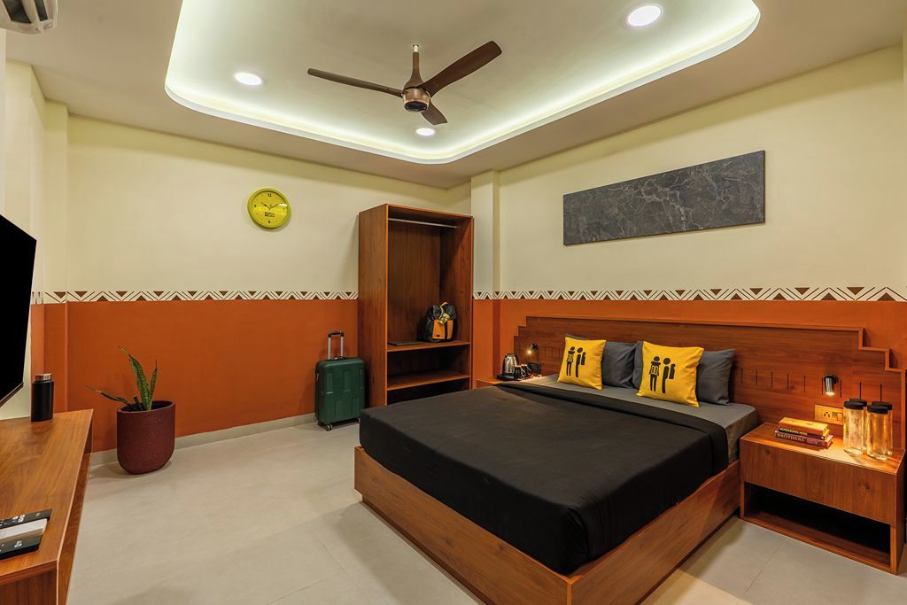 A hotel room with a bed, a wooden headboard, a nightstand, a wall clock, a ceiling fan, and a potted plant.