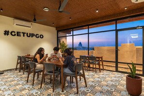 Un grupo de personas sentadas a una mesa en una habitación con un ventanal grande, techo de madera y un letrero con un hashtag en la pared.