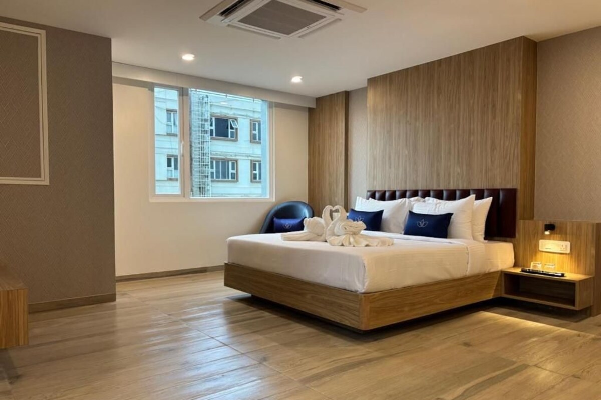 A hotel room with a large bed, a nightstand, a lamp, and a window with a view of buildings.