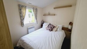 A bedroom with a bed, a window with curtains, a bedside table, and a shelf above the bed. - 3-bed Lodge, Sleeps 6 Near Beach With Indoor Pool (Hull)