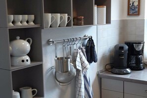 Eine Küche mit Regalen, auf denen weiße Tassen und ein Wasserkocher stehen, eine Kaffeemaschine sowie eine Kleiderstange mit einem Stoff.