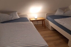 Ein Zimmer mit zwei Betten, jeweils mit gestreifter Bettwäsche, einem kleinen Holztisch mit Lampe und einem an der Wand befestigten Regal.