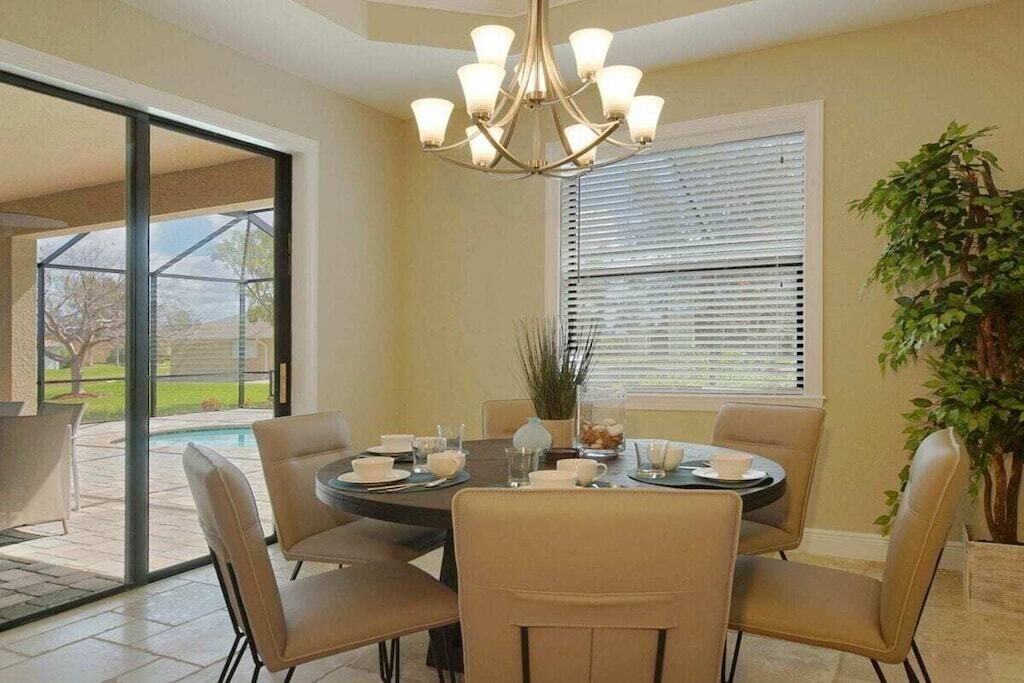 A dining area with a round table, chairs, a chandelier, and a view of the outdoors.