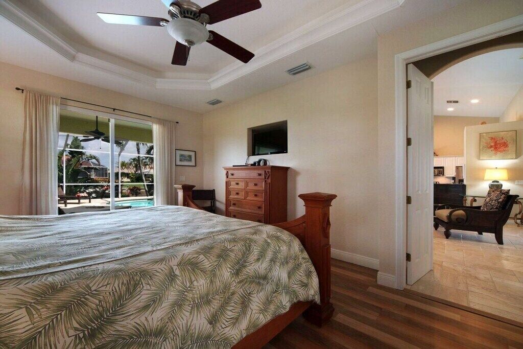 A bedroom with a bed, a ceiling fan, a wooden dresser, and a view of a pool through a sliding glass door.