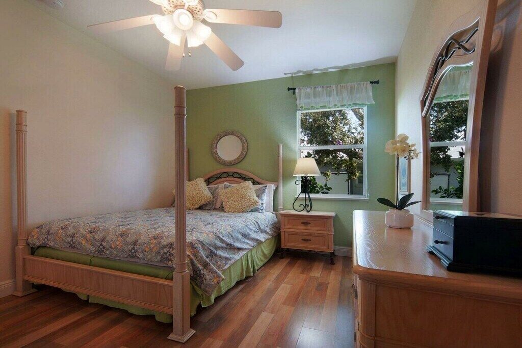 A bedroom with a four-poster bed, a nightstand, a dresser, a ceiling fan, and a window with a view of trees.