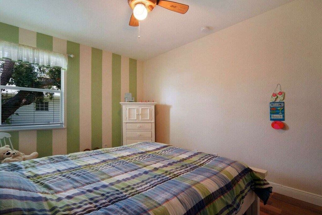 A bedroom with a bed, a dresser, a ceiling fan, a window with blinds, and a wall-mounted sign.