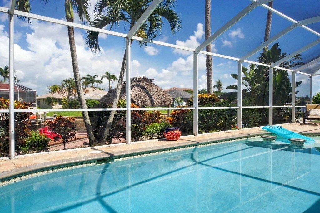 A clear blue pool with a white frame, surrounded by palm trees and a thatched-roof structure.