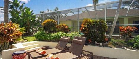An outdoor deck with lounge chairs and a table, surrounded by tropical plants and a glass-enclosed structure.