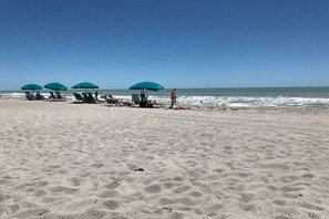 Ein Strand mit mehreren Liegestühlen und Sonnenschirmen, einem klaren Himmel und dem Meer im Hintergrund.
