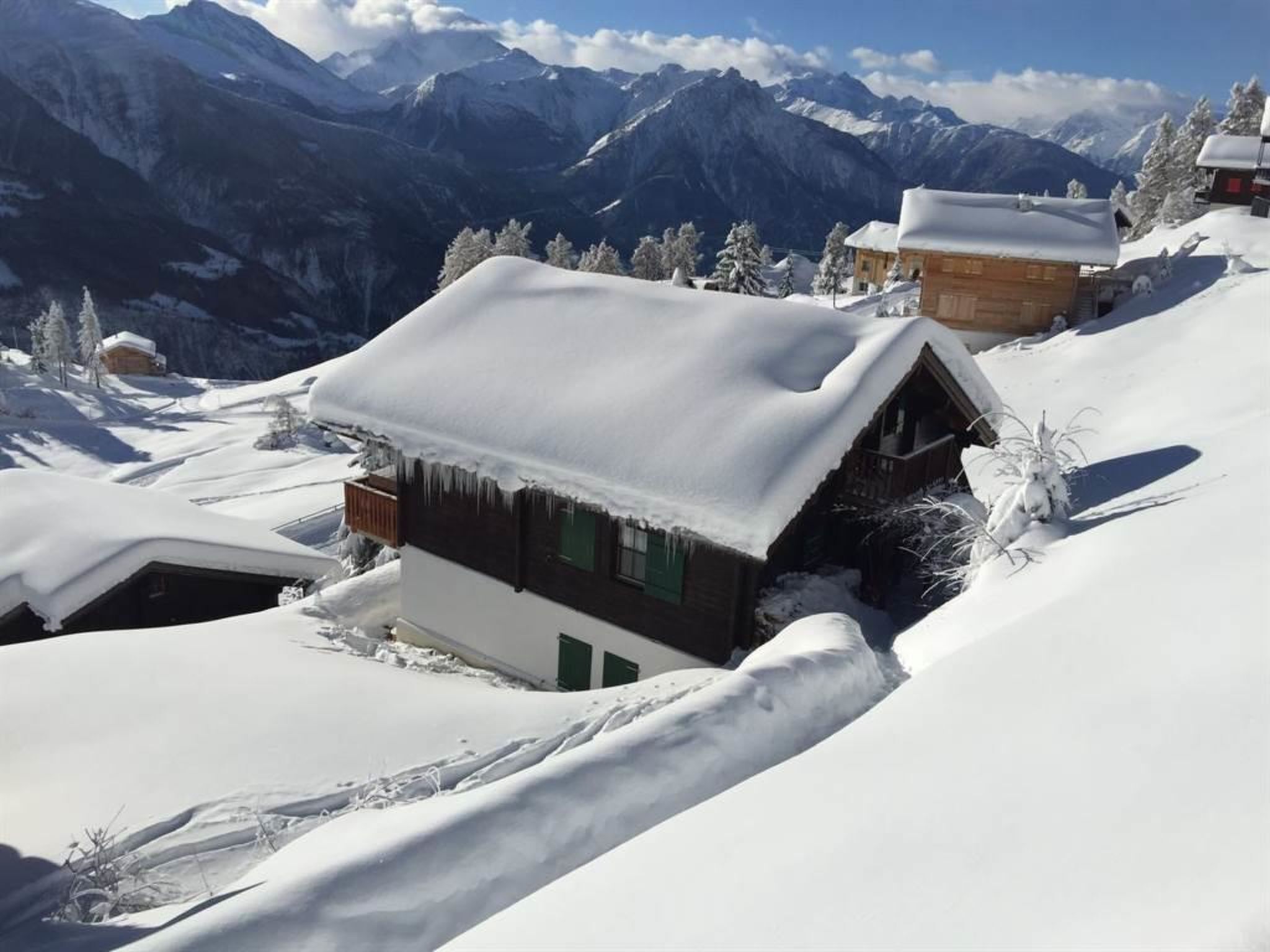 Una cabaña en la montaña cubierta de nieve, con fachada de madera, rodeada de árboles nevados y montañas al fondo.
