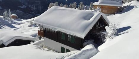 Una cabaña en la montaña cubierta de nieve, con fachada de madera, rodeada de árboles nevados y montañas al fondo.