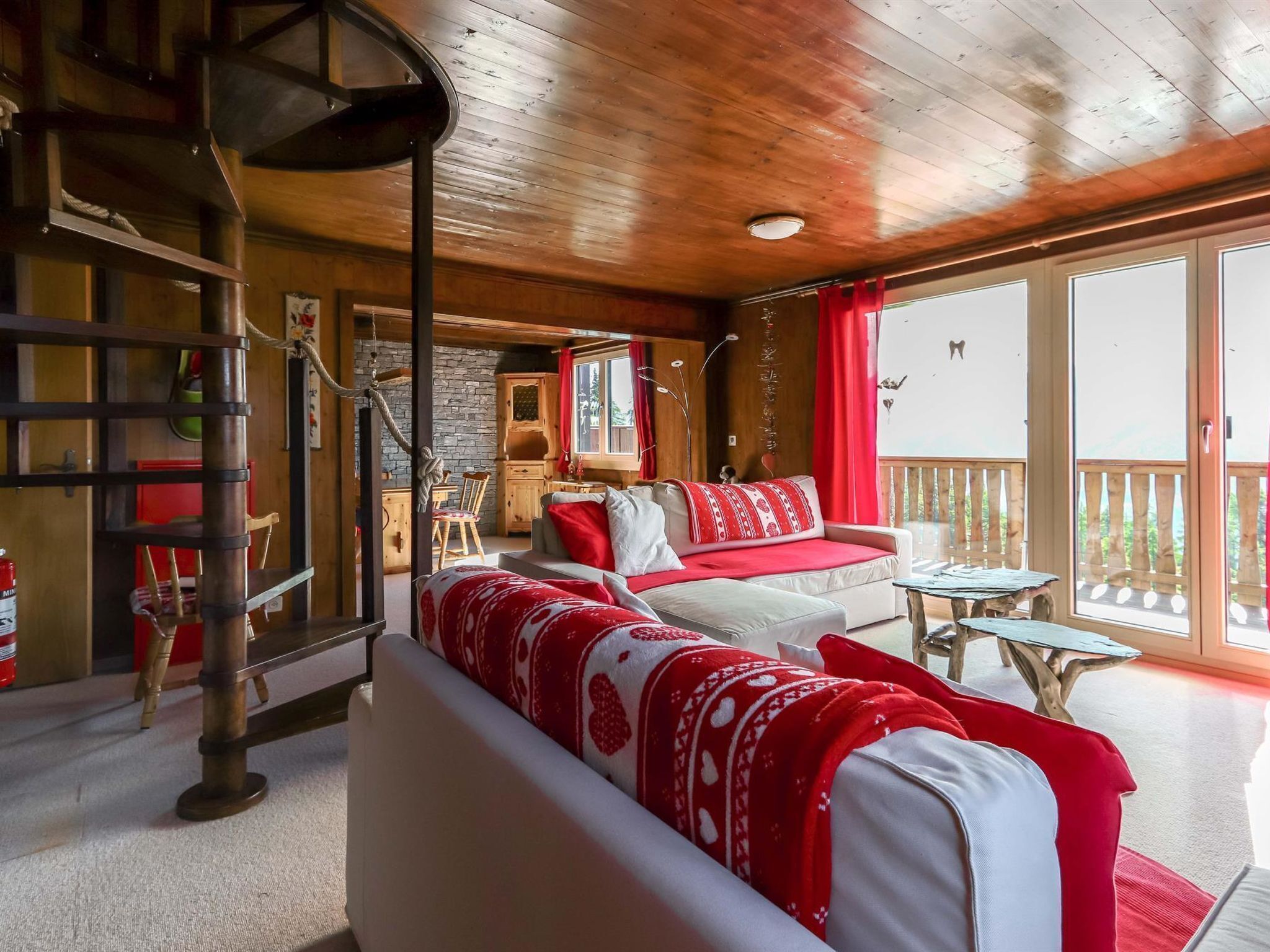 A cozy cabin interior with a wooden ceiling, a sofa with red and white bedding, a dining area with a table and chairs, and a balcony with a view.