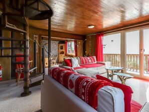 Ein gemütliches Blockhaus-Interieur mit Holzbalkendecke, einer Couch mit rot-weißer Bettwäsche, einem Essbereich mit Tisch und Stühlen und einem Balkon mit Aussicht.