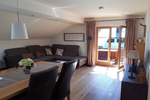 A modern living room with a sofa, dining table, chairs, and a view through a glass door to an outdoor balcony. - Holiday apartment with 1 bedroom (Bad Wiessee)