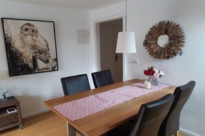Ein Essbereich mit einem Holztisch, schwarzen Stühlen, einer rosa Tischdecke, einem Wandbild einer Eule und einer Wandleuchte sowie einem dekorativen Kranz.