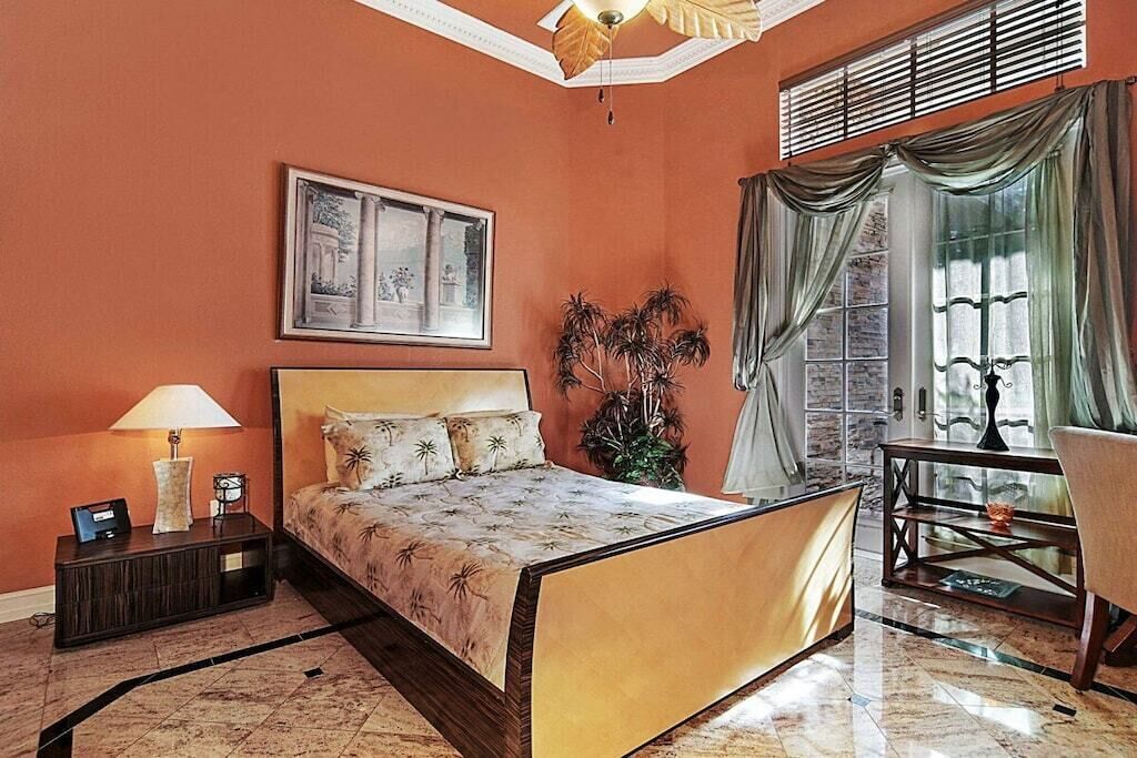 A hotel room with a bed, a nightstand, a lamp, a framed picture, a potted plant, a window with curtains, and a small table with a chair.