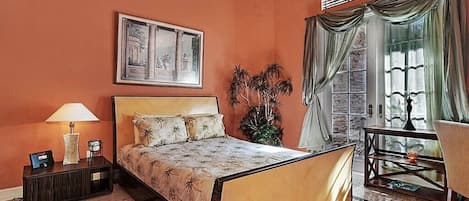 A hotel room with a bed, a nightstand, a lamp, a framed picture, a potted plant, a window with curtains, and a small table with a chair.