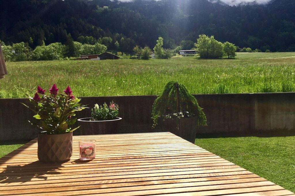 Eine Holzterrasse mit einem Blumentopf und einer Kerze mit Blick auf ein Grasfeld und einen Berg im Hintergrund.