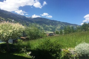 Ländliche Landschaft mit einem Haus, umgeben von Grünflächen, mit Bergen im Hintergrund.