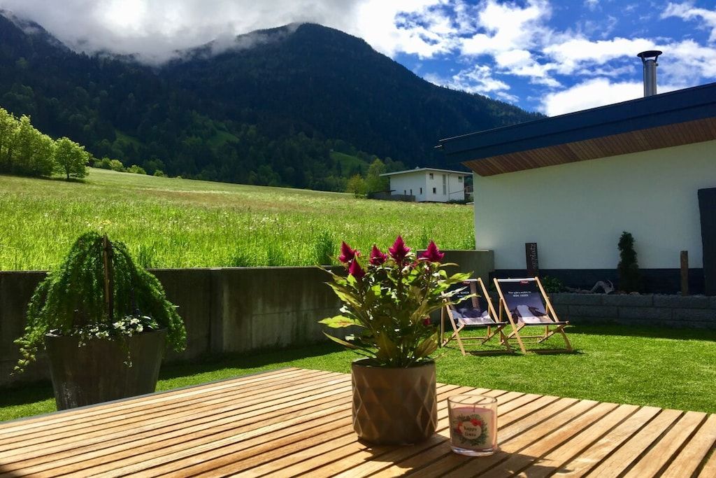 A wooden deck with a potted plant and a glass of drink, overlooking a green field and a mountain.