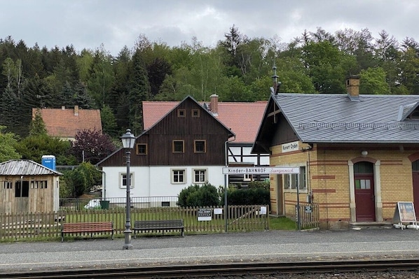 Una estación de tren con un edificio de madera, un edificio blanco y un edificio de ladrillo. Hay bancos y una valla.