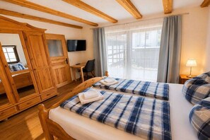Une chambre d’hôtel avec des poutres apparentes, un lit avec une couette à carreaux, un bureau avec une chaise, une télévision et une fenêtre avec des rideaux.