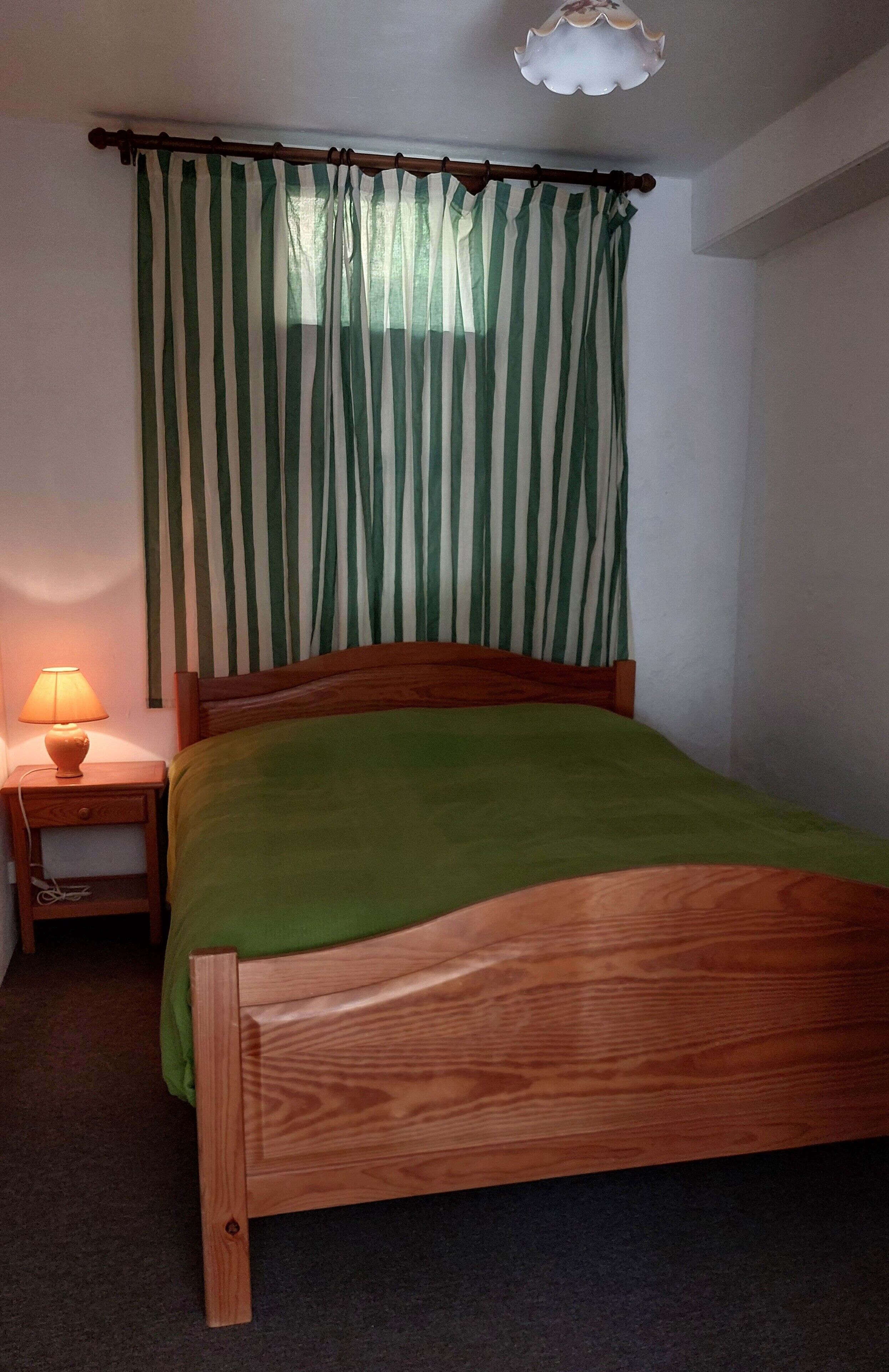 Un lit simple avec un cadre en bois et une couvre-lit verte, une table de chevet avec une lampe, et une fenêtre ornée de rideaux verts et blancs à rayures.