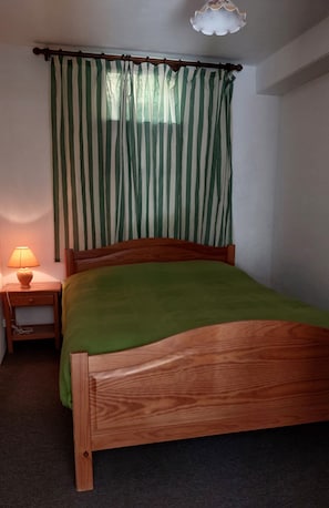 Ein Einzelbett mit Holzrahmen und einer grünen Tagesdecke, ein Nachttisch mit Lampe sowie ein Fenster mit grün-weißen Streifenstoren.