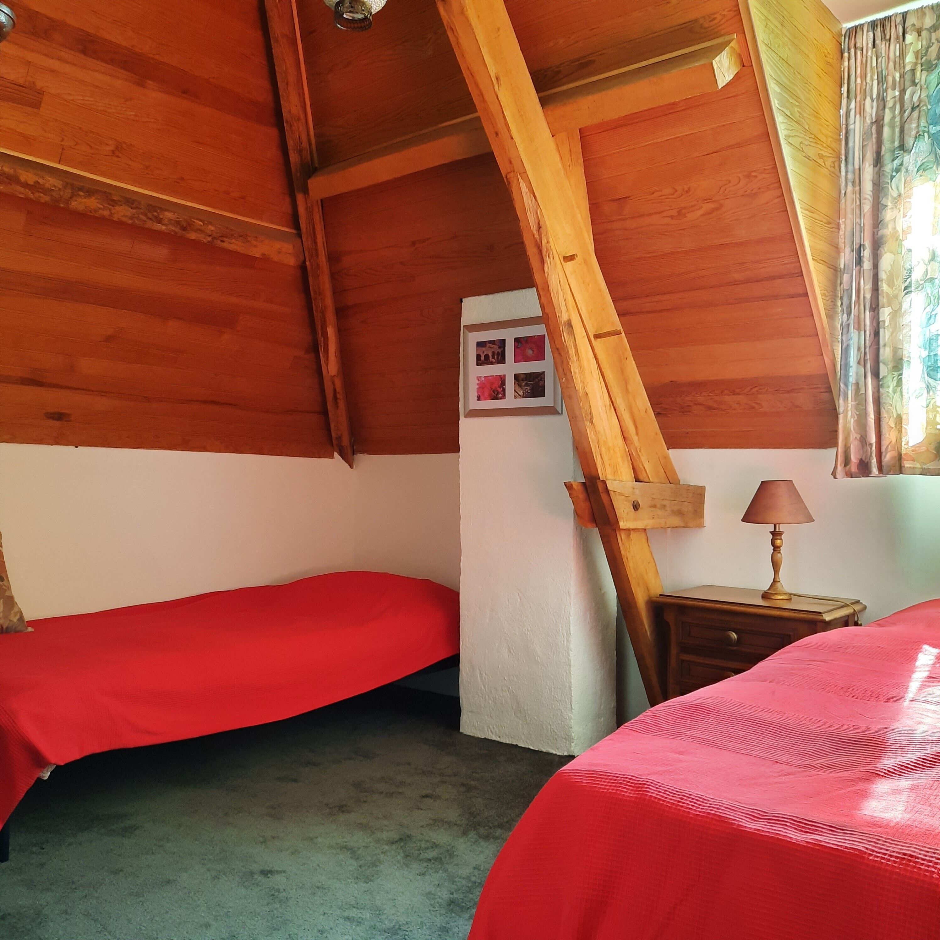 Une chambre avec deux lits, des poutres apparentes en bois au plafond, une petite table avec une lampe et une fenêtre avec des rideaux.