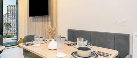 A modern dining area with a wooden table set for two, a flat-screen TV mounted on the wall, and a view of a building outside.