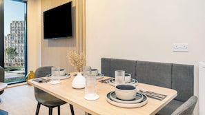 Un espace repas moderne avec une table en bois dressée pour deux personnes, un téléviseur à écran plat fixé au mur et une vue sur un bâtiment à l’extérieur.