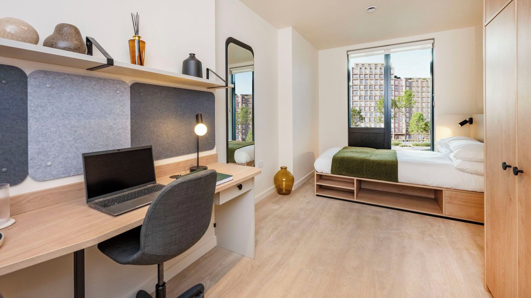 A compact bedroom with a desk, laptop, chair, bed, and a window with a view of buildings.