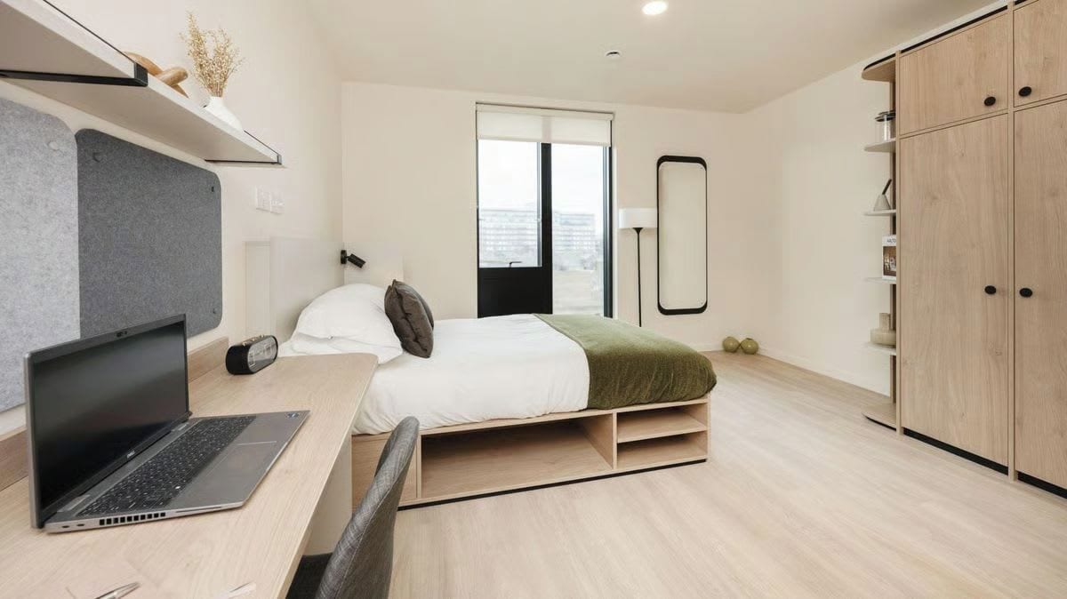 A modern bedroom with a bed, a desk with a laptop, a wardrobe, and a window with a view.